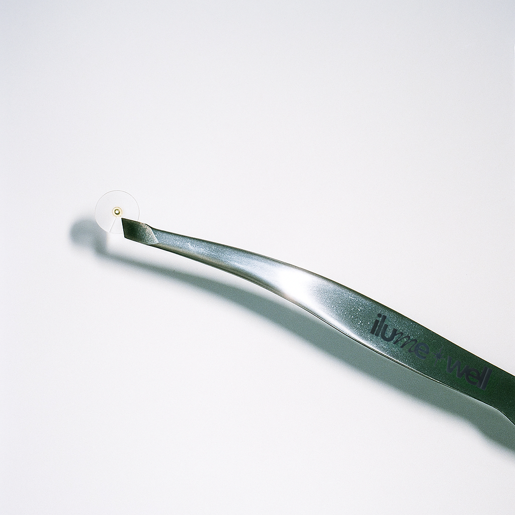 Metallic tweezer with 'ilumewell' branding, holding gold ear seeds on a white background